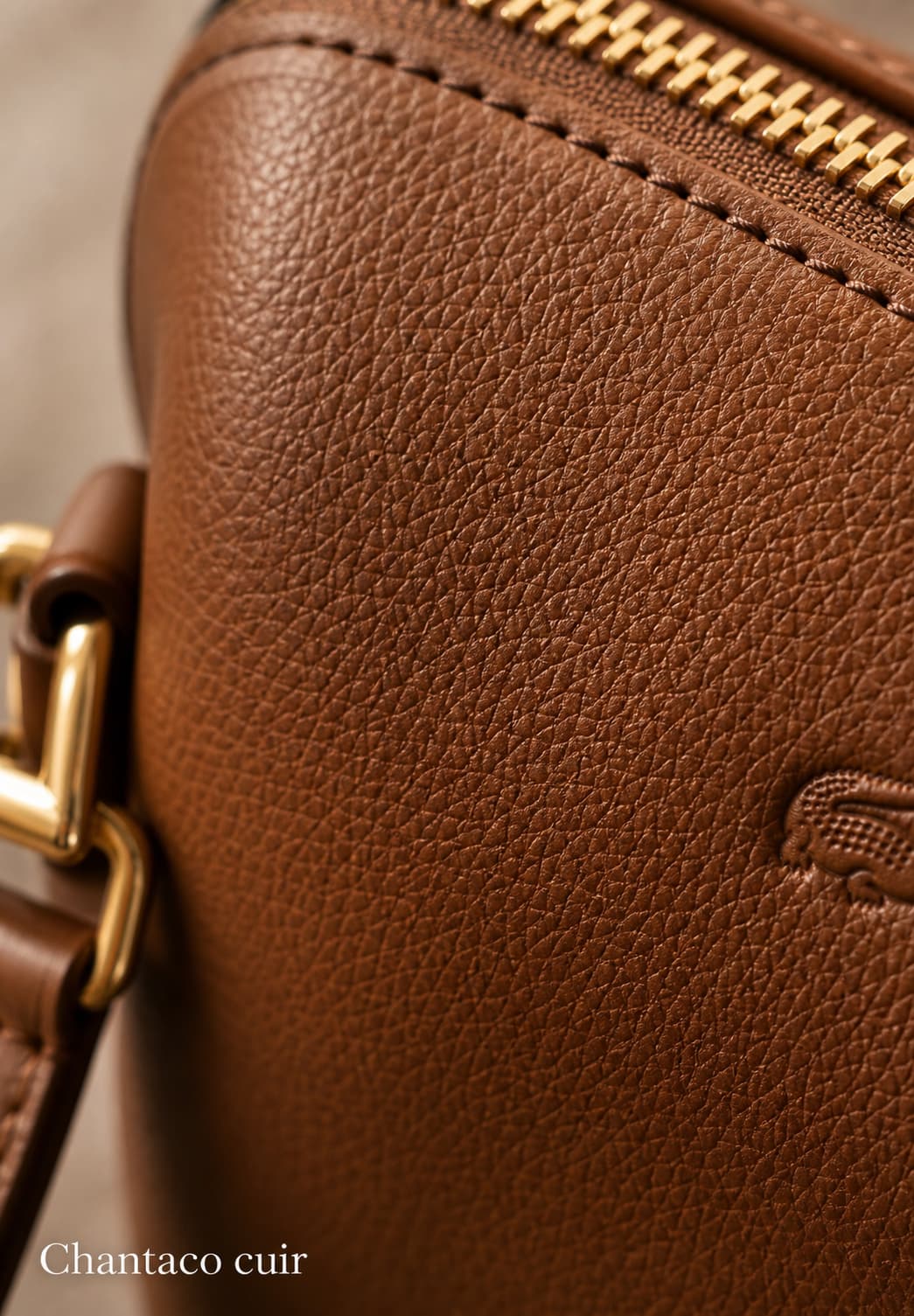 Gros plan extrême sur un sac en cuir piqué marron cognac avec texture grainée, quincaillerie en laiton et logo crocodile partiellement visibles, fond flouté et texte 