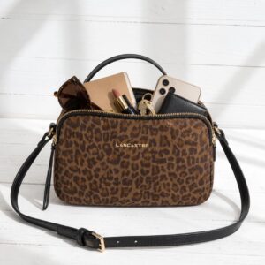 Sac besace Lancaster Jungle de couleur camel posé sur un fond en bois blanc, rempli d'objets du quotidien débordant légèrement et mettant en valeur sa contenance.