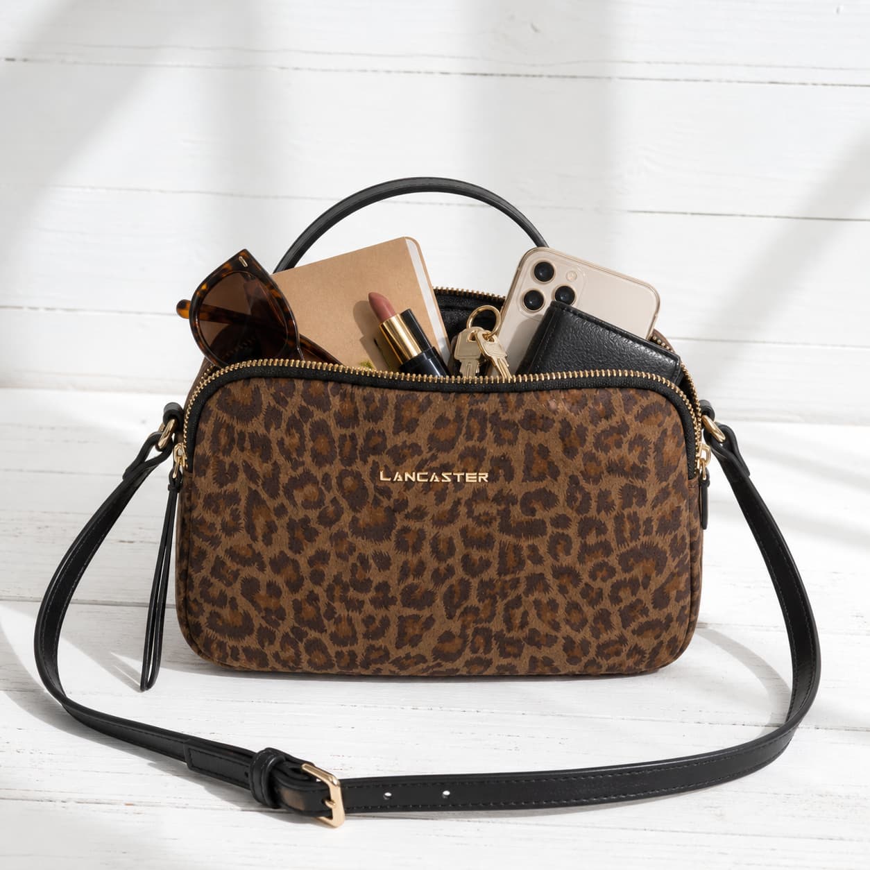Sac besace Lancaster Jungle de couleur camel posé sur un fond en bois blanc, rempli d