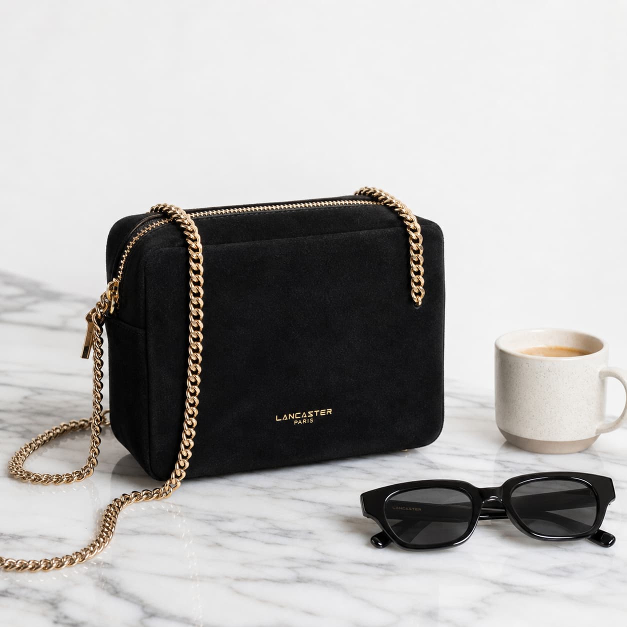 Sac Lancaster Suède Cube en suède noir avec chaîne dorée posé sur une surface en marbre à côté d