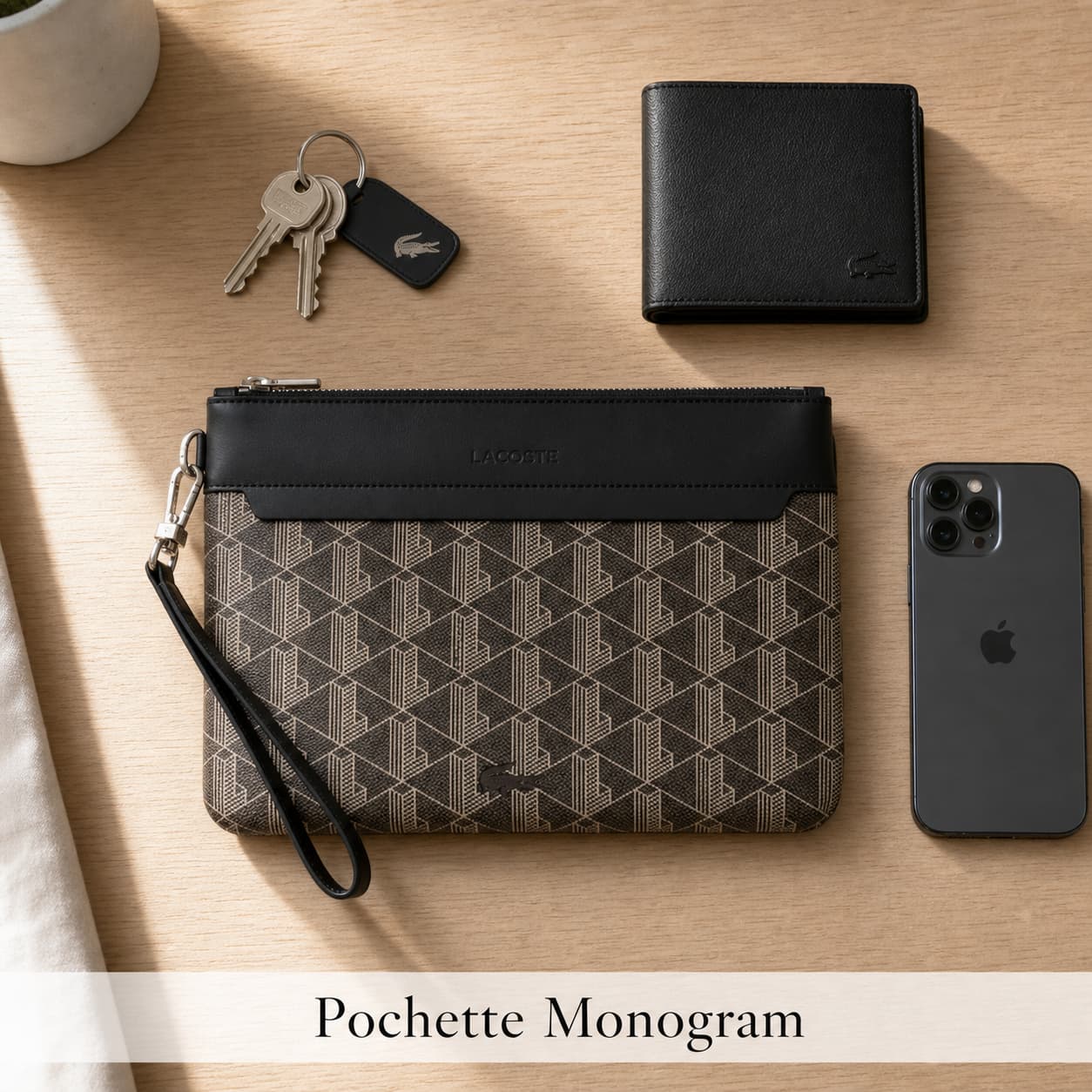 Pochette Lacoste Monogram beige et marron avec bordure noire, posée à plat sur une table en chêne clair, entourée de clés, portefeuille et téléphone, éclairage naturel avec ombres subtiles, texte 