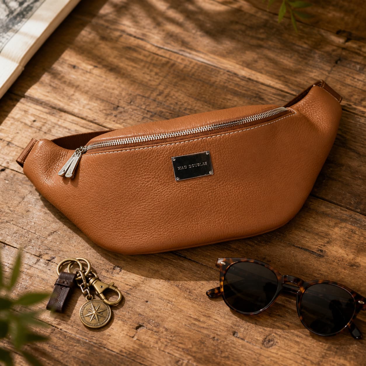Sac banane en cuir marron Mac Douglas Barbade CIENNA disposé à plat avec des lunettes de soleil et un porte-clés sur une table en bois rustique, sous la lumière naturelle du soleil, ambiance voyage tendance.
