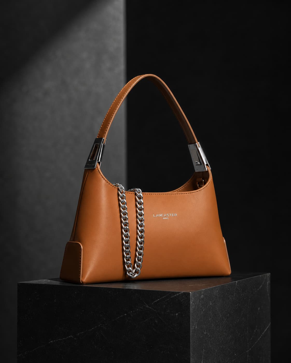 Sac Lancaster Suave Ace avec détail de chaîne, posé sur un piédestal sous un éclairage dramatique, créant une ambiance moderne et audacieuse.