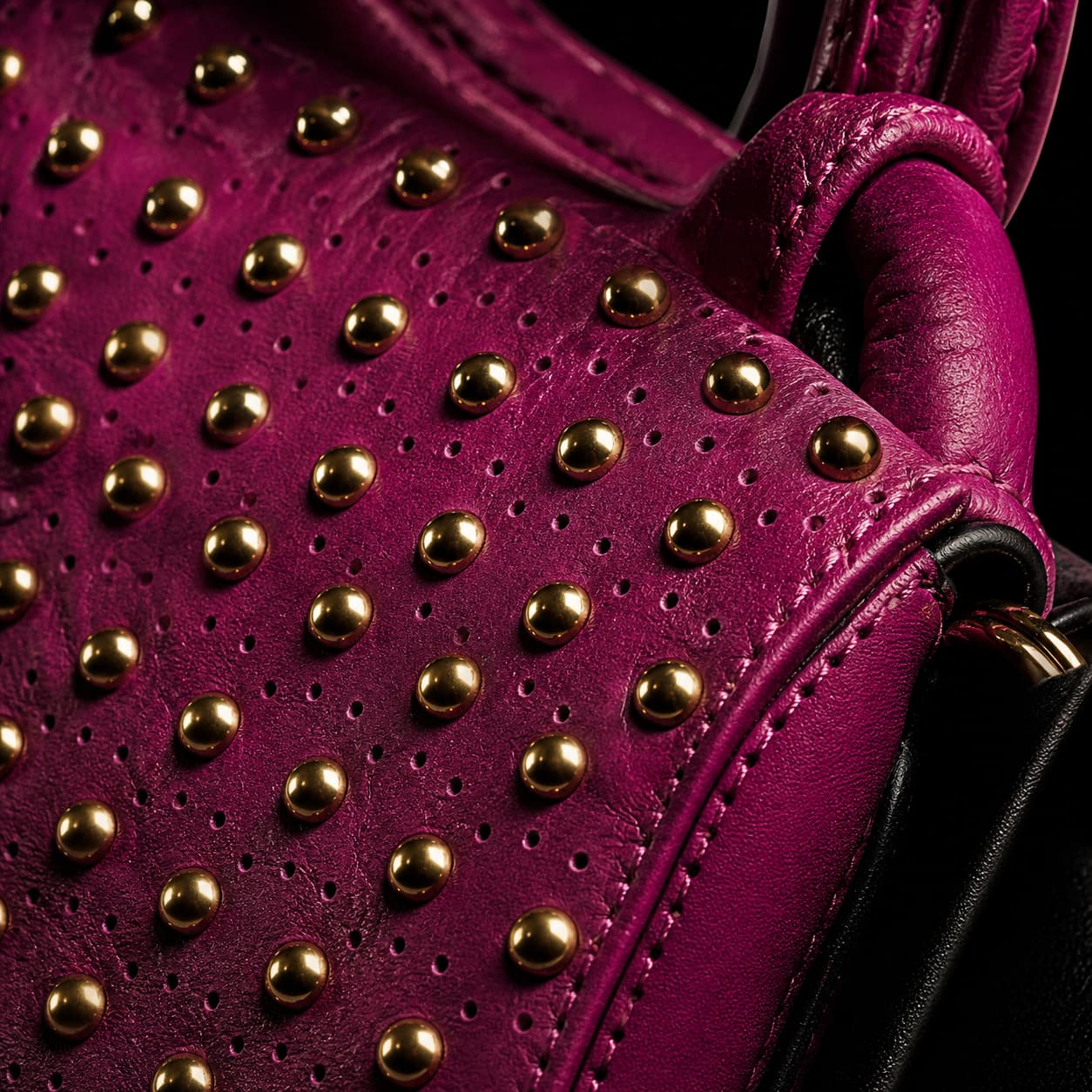 Gros plan en macro sur un sac à main Fuchsia Paris en cuir noir, orné de clous dorés et de mini-perforations, éclairé par une lumière latérale dramatique sur fond sombre.