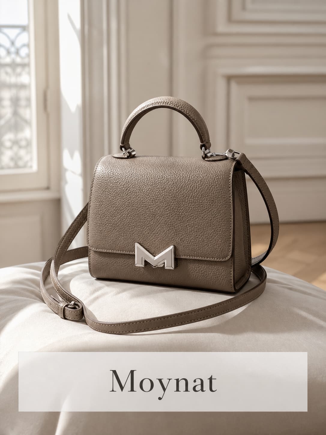 Sac Moynat Réjane en cuir taupe avec fermoir M, posé sur un coussin en soie dans un appartement parisien aux moulures classiques, lumière douce de fenêtre, avec le nom